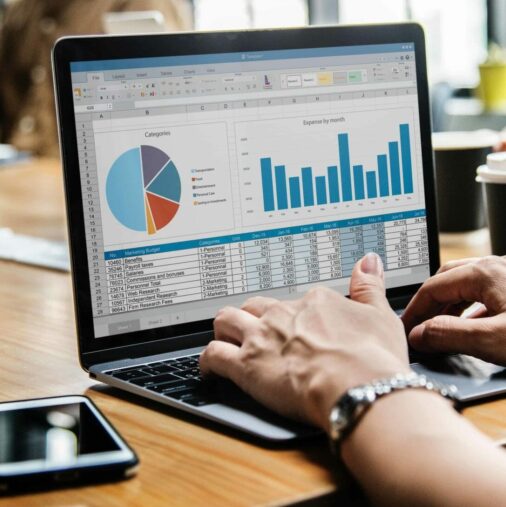 A person works on a laptop displaying spreadsheets with a pie chart and bar graph, while others sit around a table with papers, coffee cups, and a smartphone.
