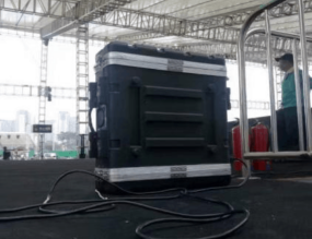 A large black equipment case with cables attached sits on a stage under a tent. Metal railings and a person in the background are visible. A large black equipment case with cables attached sits on a stage under a tent. Metal railings and a person in the background are visible.