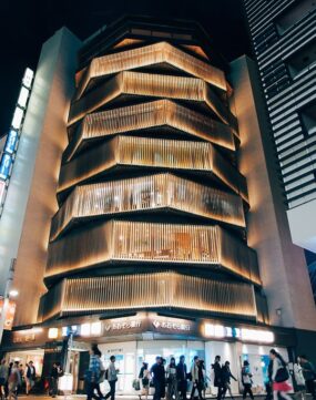 A modern, multi-story building with illuminated, layered wooden slats on the facade at night; people walk by on the street below. A modern, multi-story building with illuminated, layered wooden slats on the facade at night; people walk by on the street below.