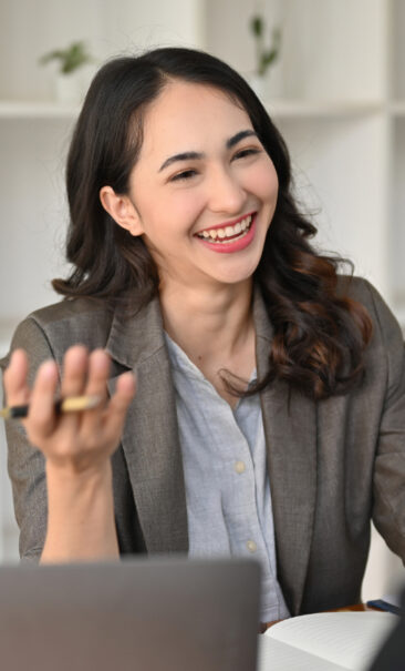 A woman in a blazer sits at a desk, smiling and gesturing with a pen in hand, with a notebook and laptop in front of her. A woman in a blazer sits at a desk, smiling and gesturing with a pen in hand, with a notebook and laptop in front of her.