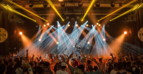 A large crowd with raised hands faces a stage with bright spotlights and a performer, in an indoor concert or music festival setting. A large crowd with raised hands faces a stage with bright spotlights and a performer, in an indoor concert or music festival setting.